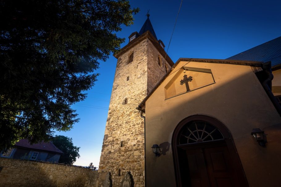 Leuchtender Kirchturm mit Kreuz und blauem Abendhimmel im Hintergrund.