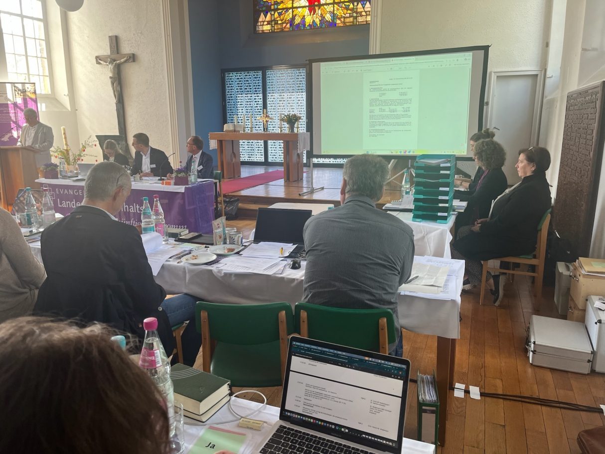 Sitzung in einer Kirche mit Menschen, Tischressourcen und Präsentation im Hintergrund.