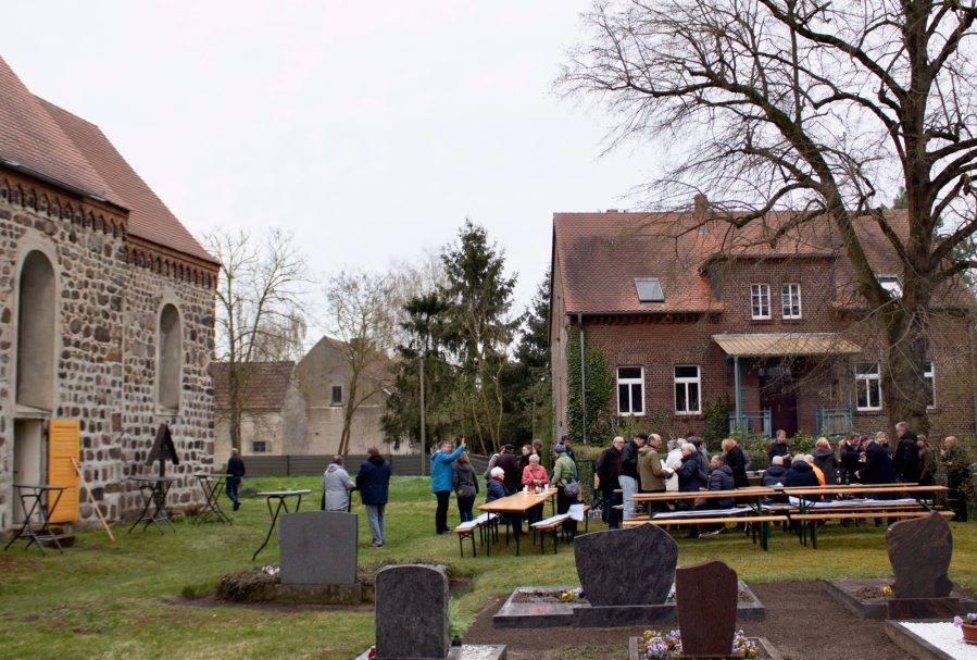 Eine Gruppe von Menschen versammelt sich im Freien neben einer Kirche und einem Friedhof.