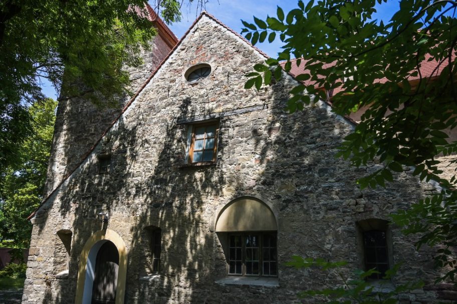 Steinerne Wand mit Fenster, umgeben von Pflanzen und Schatten, unter blauem Himmel.