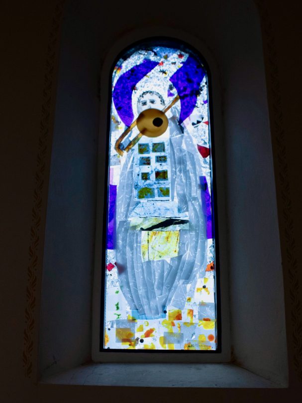 Buntes Glasfenster mit einer Figur, die eine Trompete hält und musikalische Noten zeigt.