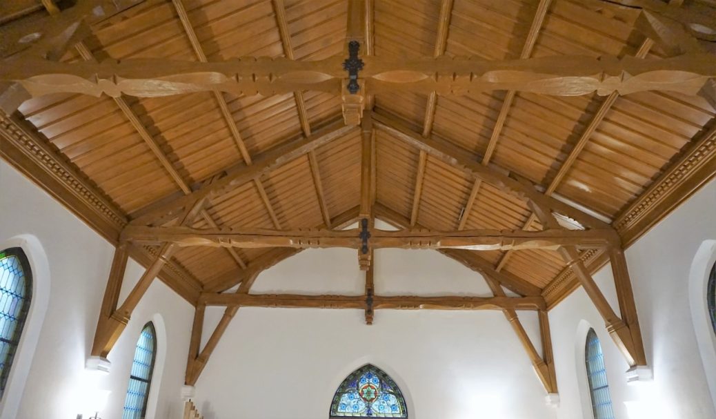 Holzdecke einer Kirche mit sichtbaren Balken und ovalen Fenstern an den Wänden.