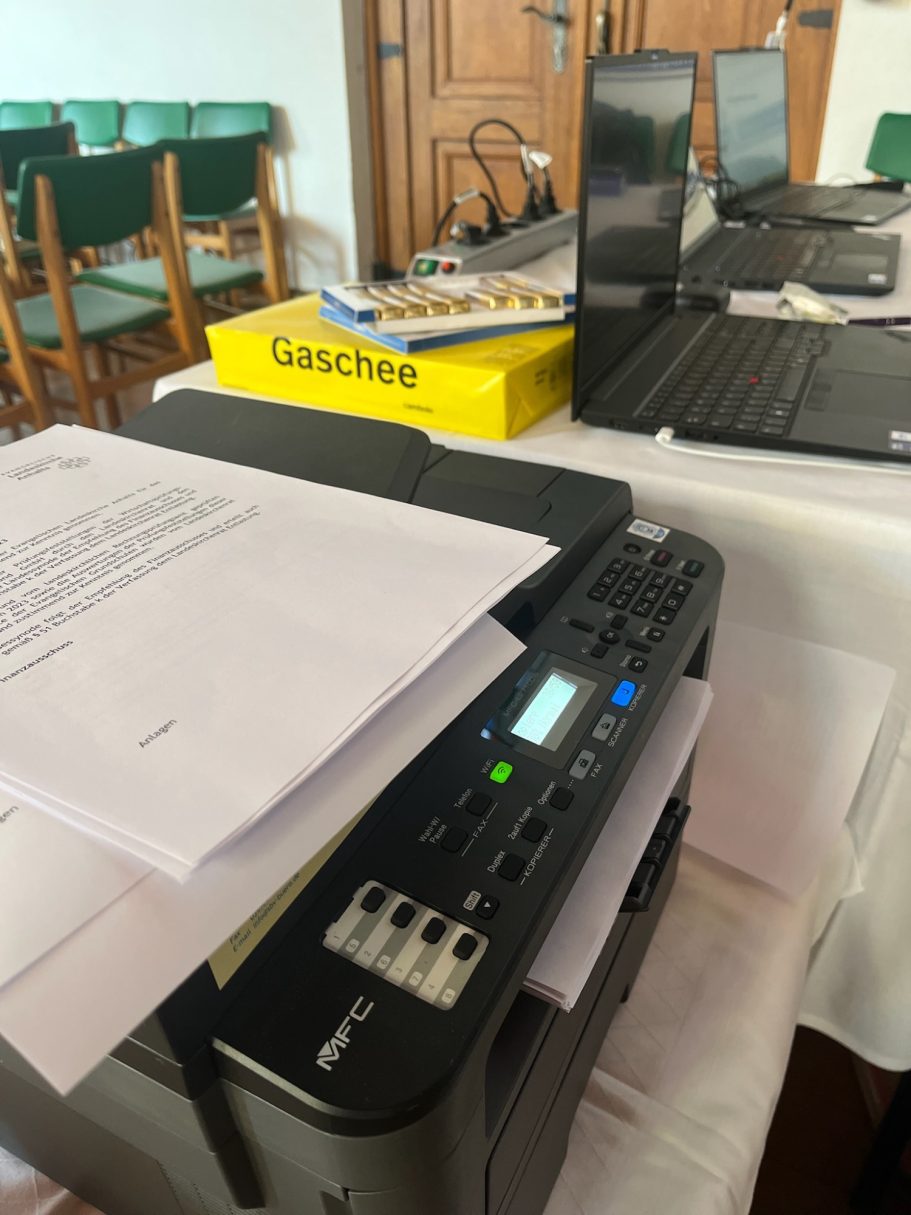 Drucker mit ausgedrucktem Papier und Laptop im Hintergrund auf einem Tisch.