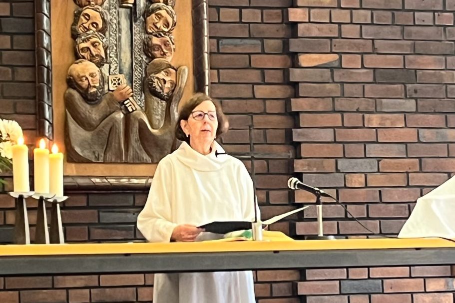 Frau mit Kreuz und Tisch, im Hintergrund Musiker mit Instrumenten.