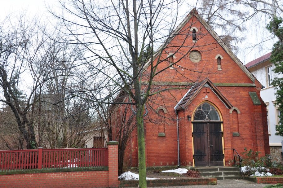 Rote Backsteinkirche mit Holzportal, umgeben von kahlen Bäumen und einem Zaun.