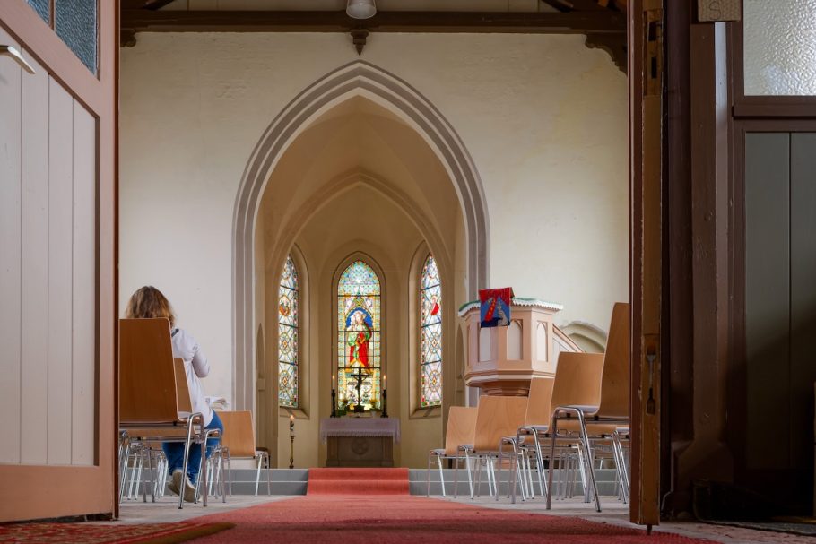 Blick durch eine Kirchentür auf den Altar mit farbigen Fenstern und Holzstühlen.