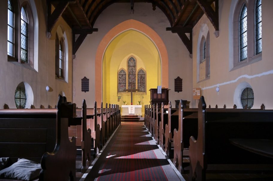 Innenansicht einer Kirche mit Holzbänken und einem beleuchteten Altarbereich.
