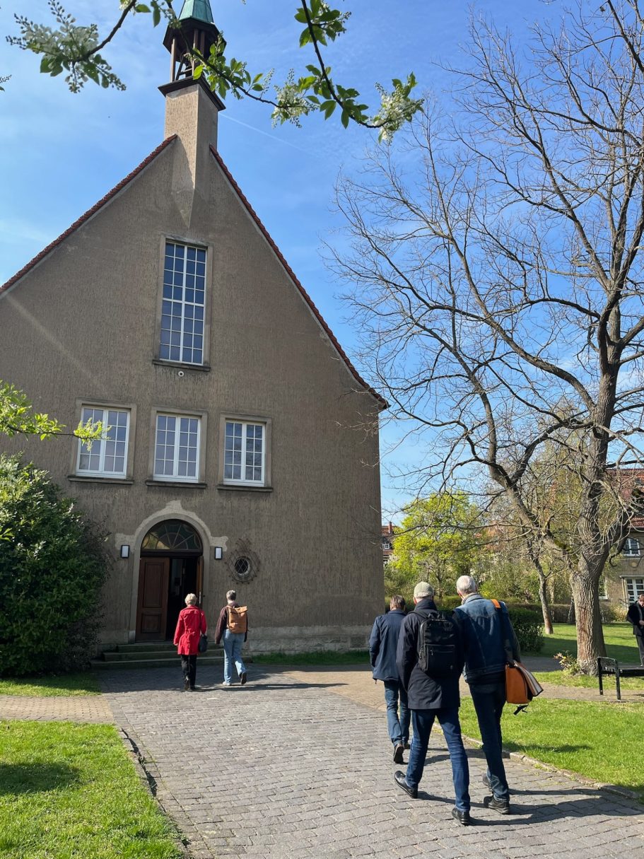 Gruppierung von Personen, die auf einen Eingang einer Kirche zulehnen, umgeben von Bäumen.
