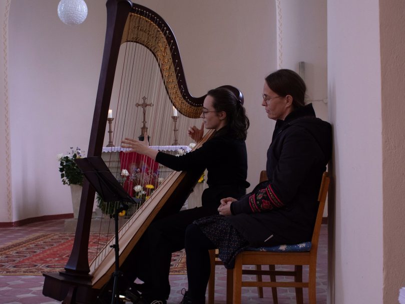 Frau spielt Harfe in einem Kirchenraum, Mann sitzt daneben und beobachtet.