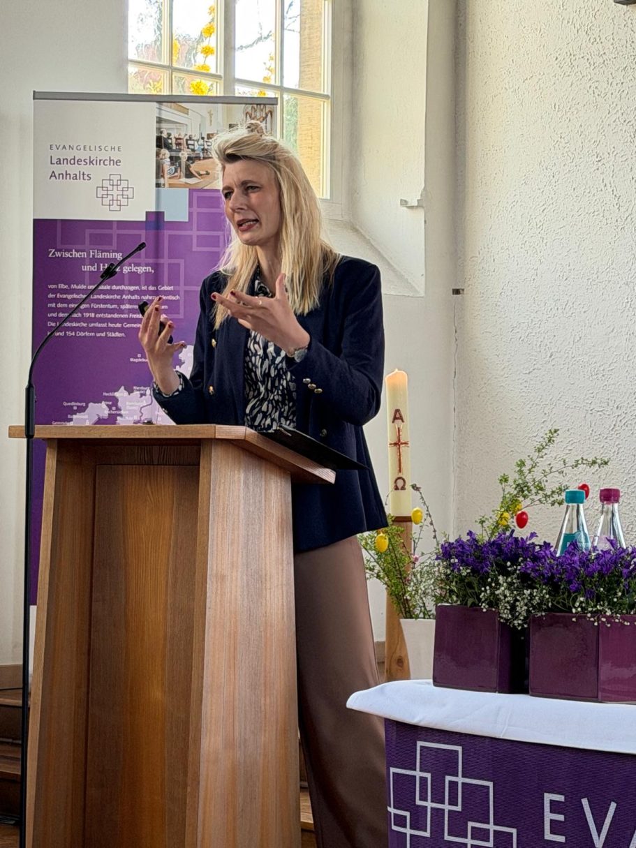 Frau spricht am Rednerpult, im Hintergrund Blumen und eine Präsentation sichtbar.