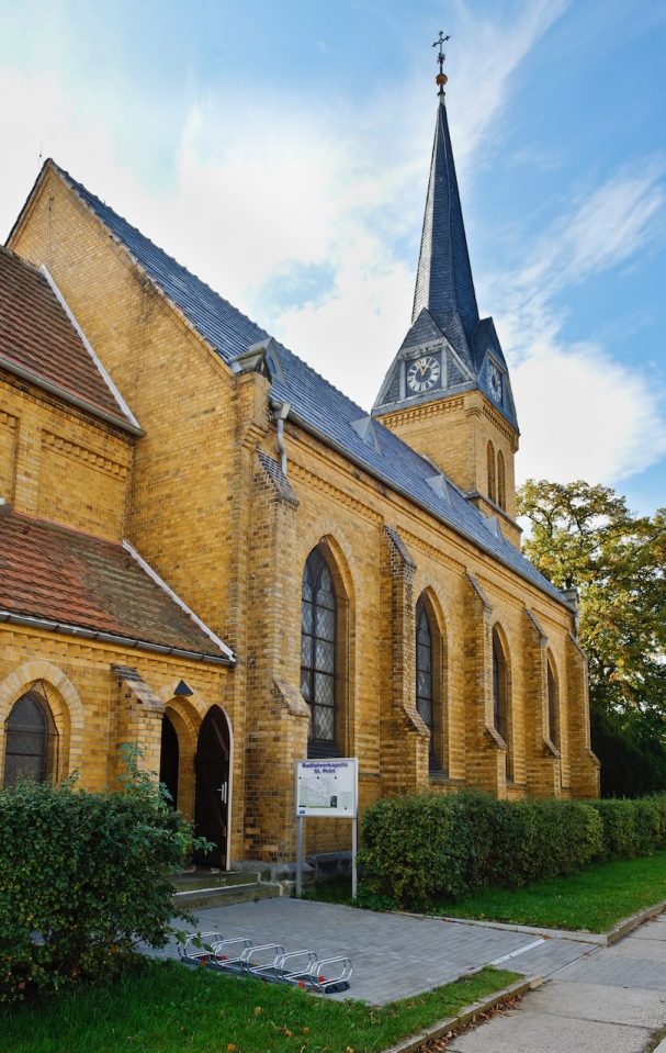 Fachwerk-Kirche mit spitzem Turm, umgeben von Bäumen und einer grünen Wiese.