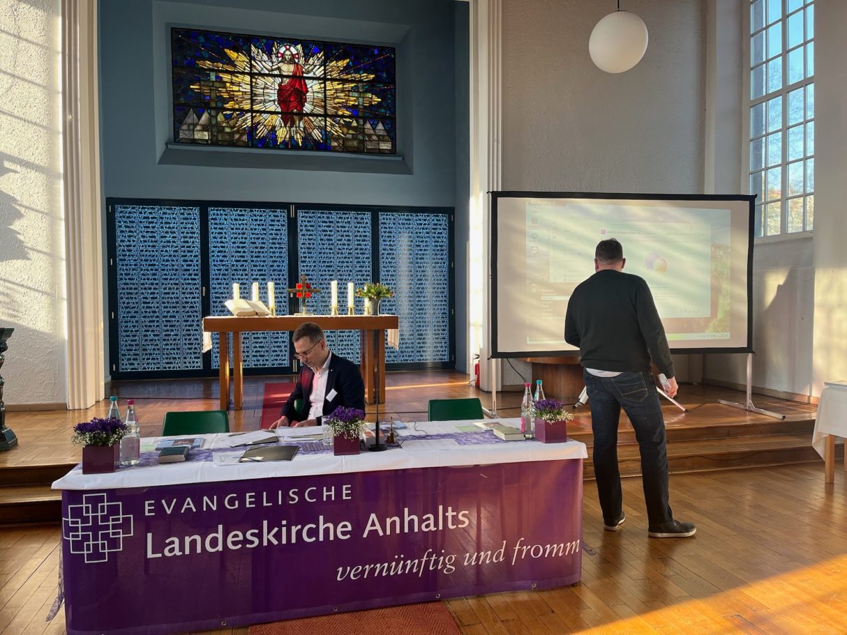 Ein Raum mit einem Tisch, einem Bildschirm und einem Mann, der auf eine Präsentation schaut.