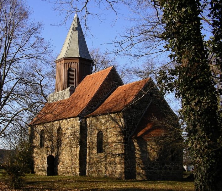 Historische Steinkirche mit spitzem Dach und umgebenden Bäumen.