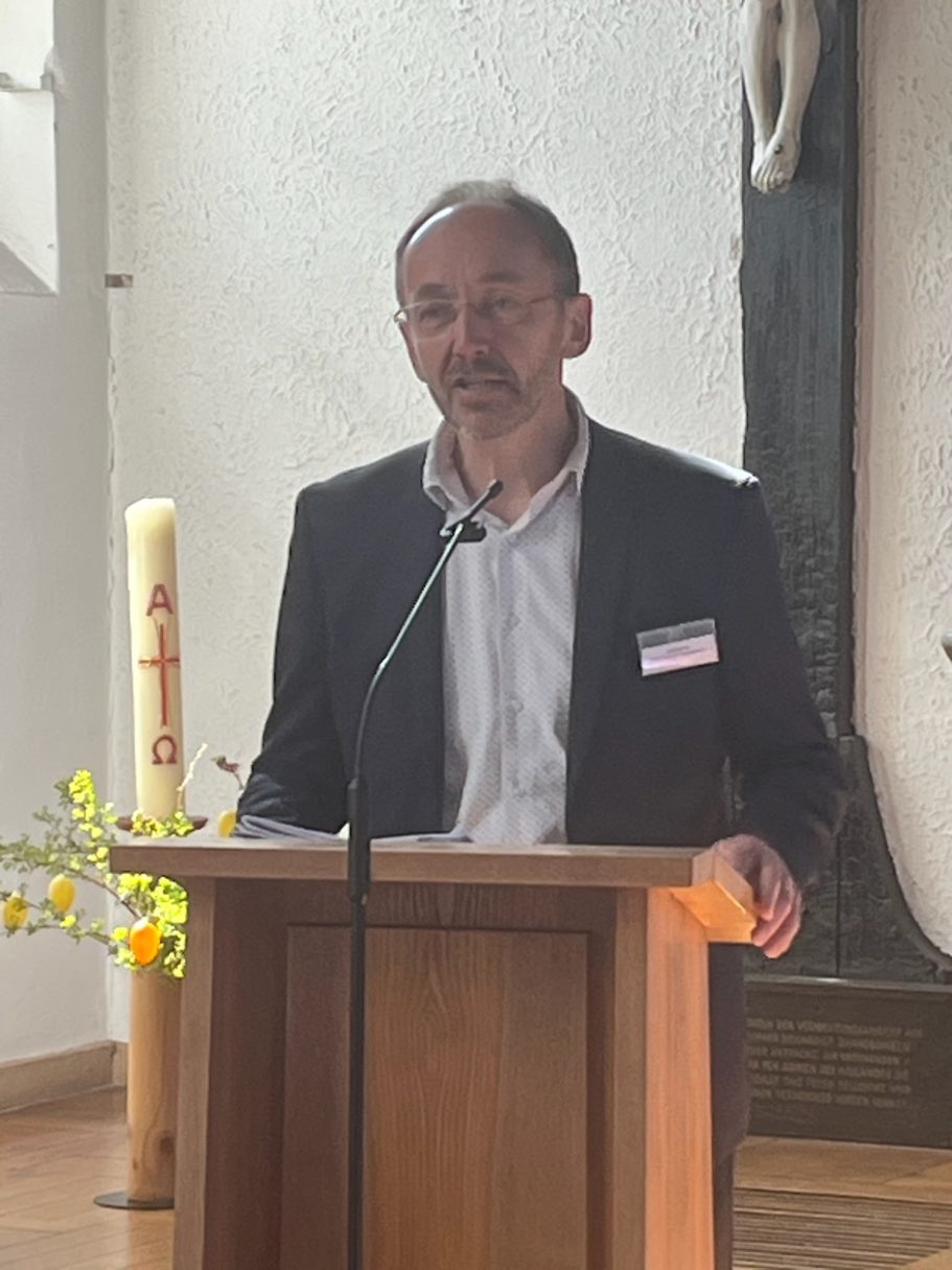 Mann spricht an einem Podium, im Hintergrund eine Kerze und eine Pflanzenanordnung.