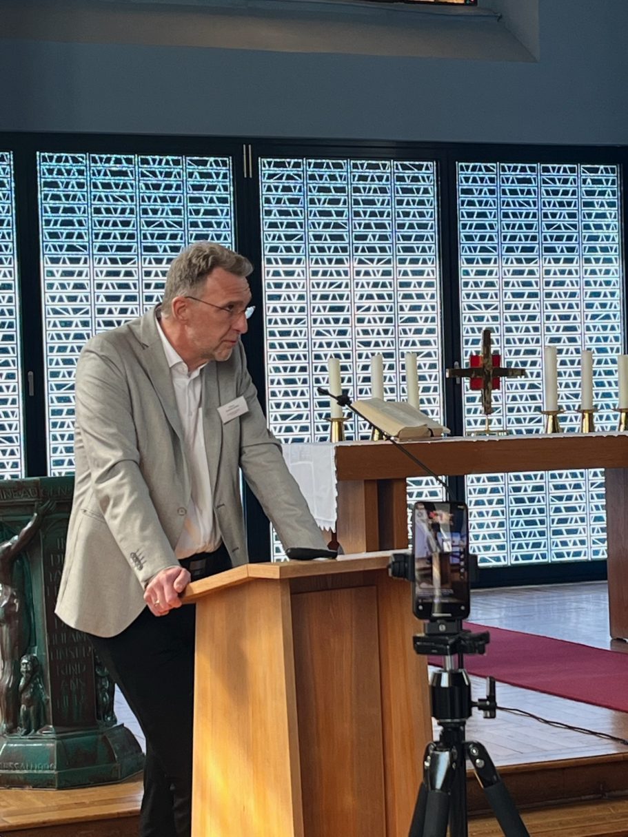 Ein Mann spricht an einem Rednerpult in einem kirchlichen Raum mit Kerzen und einem Kreuz.