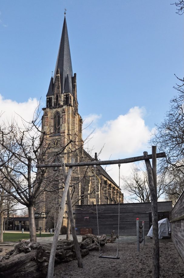 Eine Kirche mit spitzem Turm, im Hintergrund ein Spielplatz mit Schaukel.