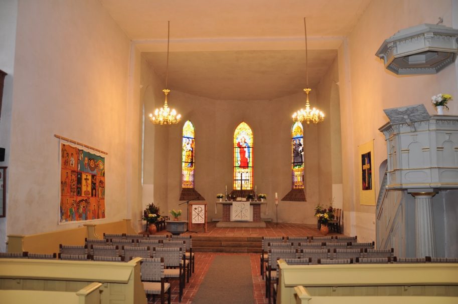 Innenansicht einer Kirche mit bunten Fenstern, einem Altar und Kirchenbänken.
