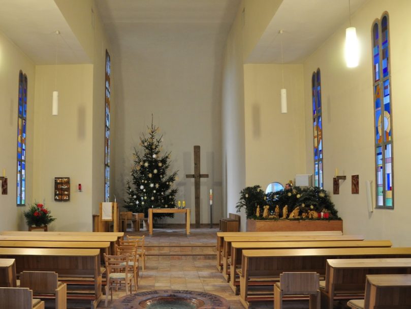 Helle Kirche mit einem Weihnachtsbaum, Holzbänken und bunten Fenstern.