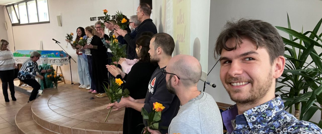 Menschen bilden einen Kreis auf einer Wiese, umgeben von Bäumen und Gebäuden.