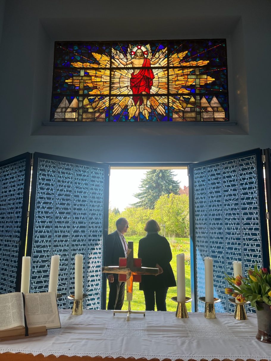 Blick auf zwei Personen, die vor einer offenen Tür mit einem Kirchenfenster stehen.